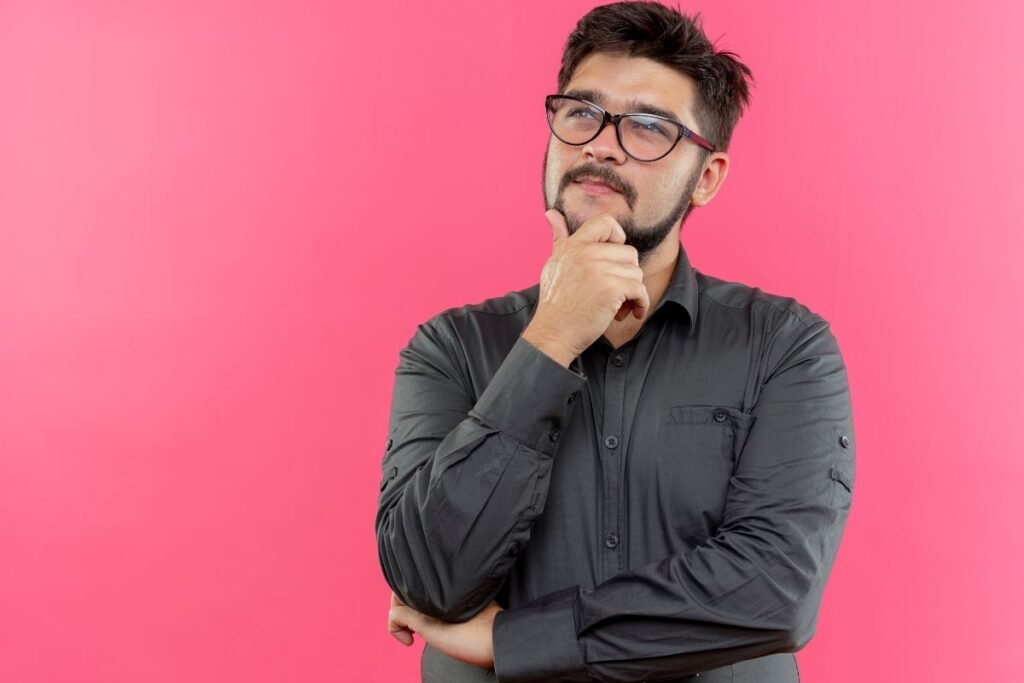 Looking up thinking young BIT An aspirant wearing glasses putting hand on chin, isolated on a pink wall