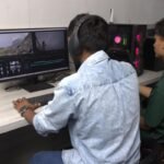 Two boy students—one in a blue shirt and another in a green t-shirt—editing on a gaming PC, shot taken from their back.